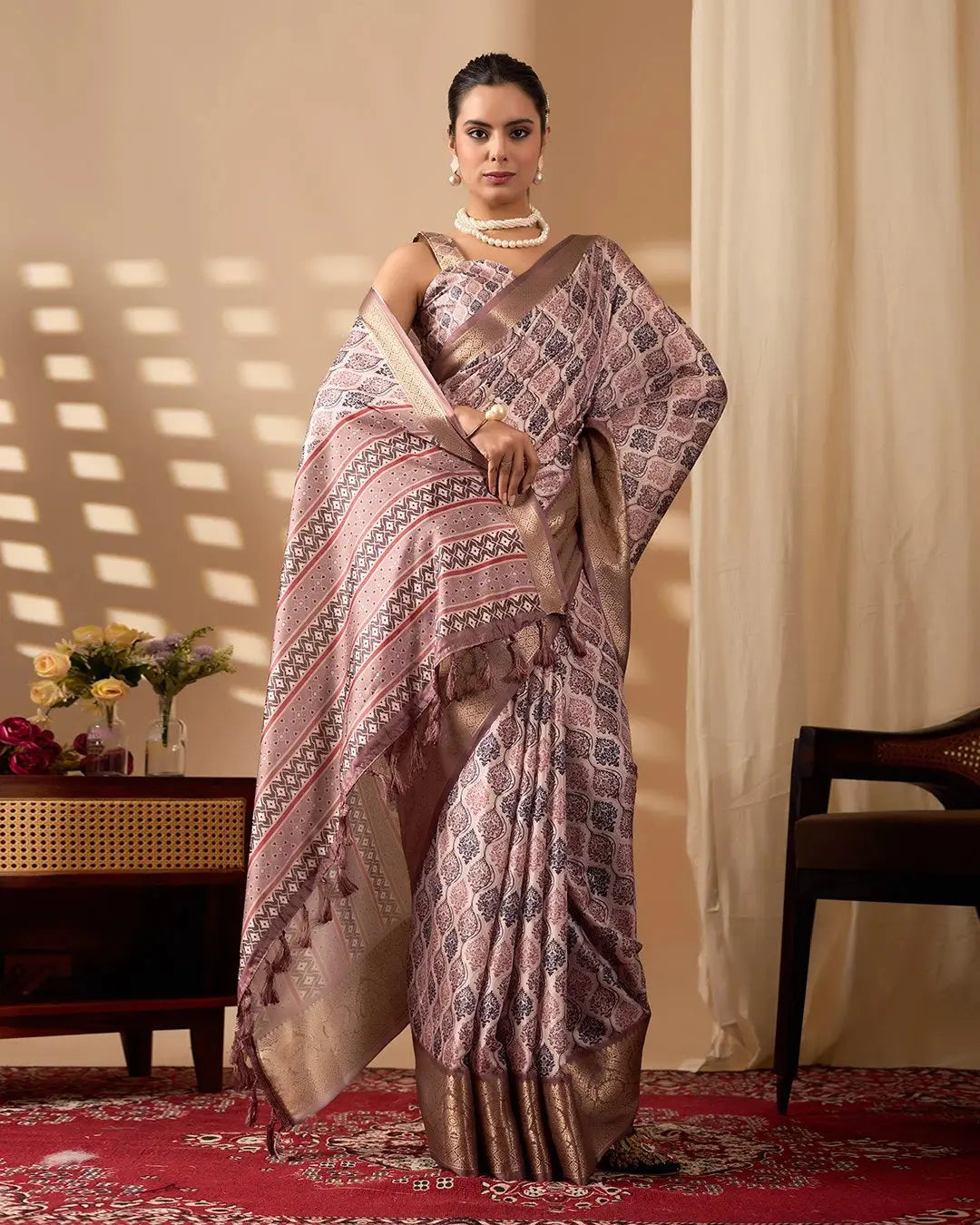 Woman wearing a patterned saree in a room with a table and flowers.